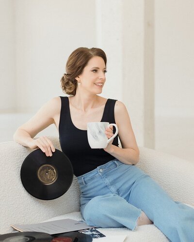 Woman with light skin and dark hair is stilling on a white couch holding a vinyl album in one hand and cup of coffee in the other. Looking off to side and smiling, wearing black tank and jeans.