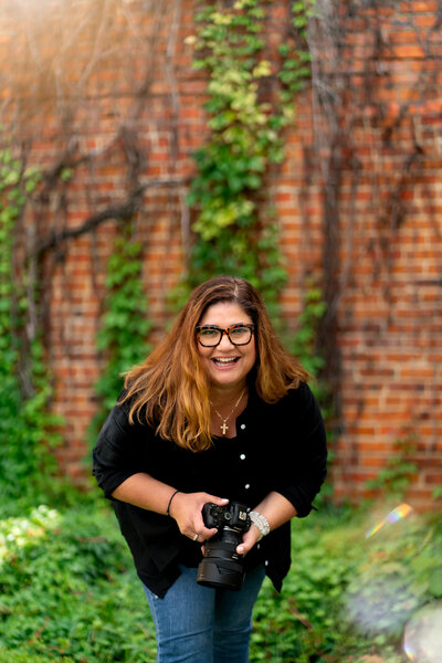 Liza Hondros holding her camera and smiling for a photo with a greenery and brick backdrop