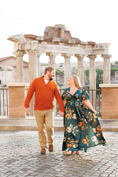 A couple walking in the roman forum at golden hour with the wife wearing a flow green dress. Taken by Rome Photographer, Tricia Anne Photography.