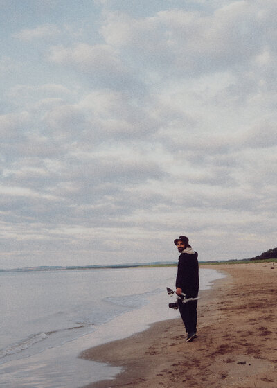 A person stands alone on a quiet beach holding a camera, looking out at the water under a soft, overcast sky for a calm, cinematic feel.