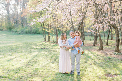 A mother, father, and their two young children stand near some cherry blossom trees at the National Arboretum during their family photo session