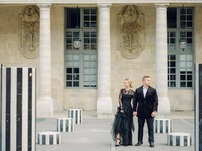 a man and woman in a black suit and black tulle dress standing shoulder to shoulder together and looking off into different directions  at palais royal in  Paris