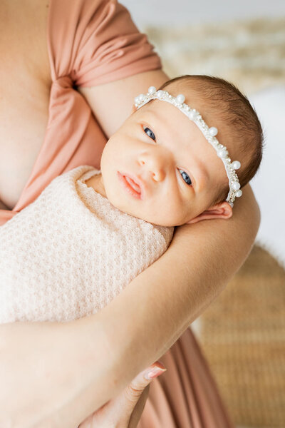 Mom in a peach dress holding her newborn wrapped in a cream blanket and pearl headband