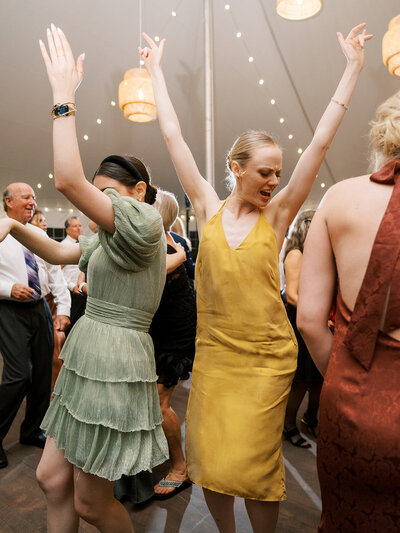 Guests dancing on the dance floor at the wedding reception with their arms in the air and colorful dresses on.