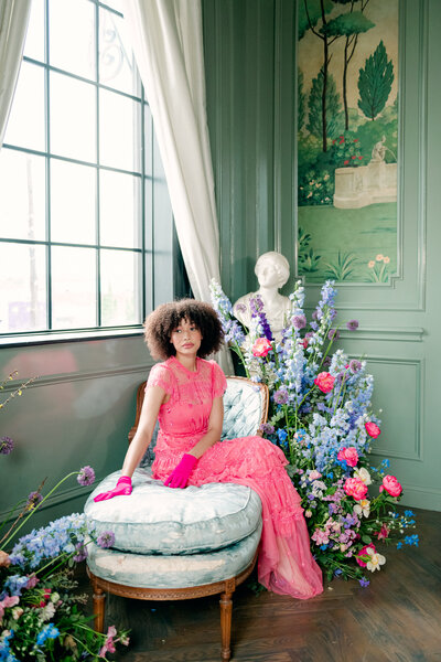 Woman in pink jumpsuit sitting on white chaise with florals surrounding her, Dallas wedding photographer
