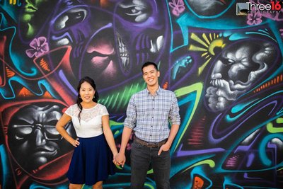Engaged couple pose holding hands during photo session in front of a colorful mural on a wall in the LA Arts District