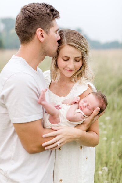 Man kisses wife as he holds newborn child in an Edina field.