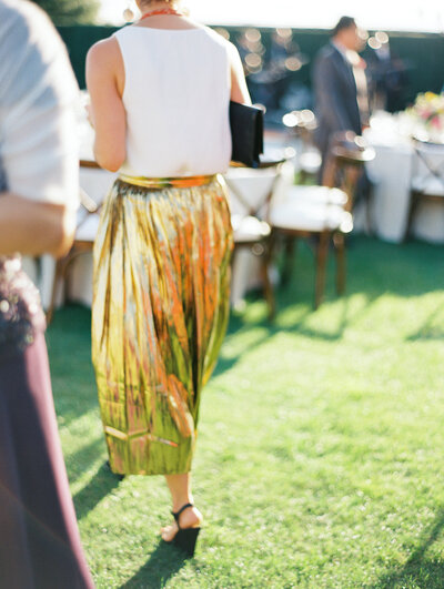 Action photo taken of a wedding guest walking by, they  wear a white shirt with long gold straight skirt. The focus of the photo is her skirt.