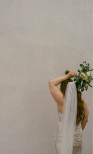 Bride with bouquet behind her back against a wall.