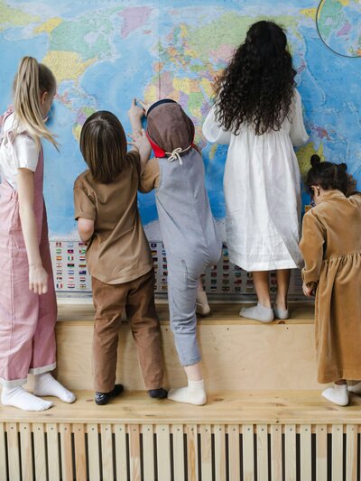 A group of children standing on a wooden platform are enthusiastically pointing and looking at different locations on a large world map displayed on the wall.