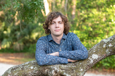 High school senior boy in a long sleeve blue patterned shirt leaning on a low hanging tree branch.