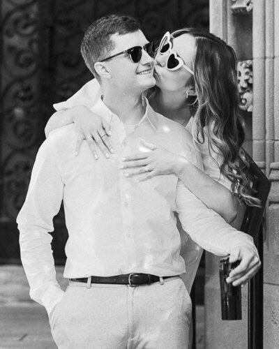 Chic Engaged couple  wearing sunglasses - the girl hugging the boy from behind and kissing him on the cheek.