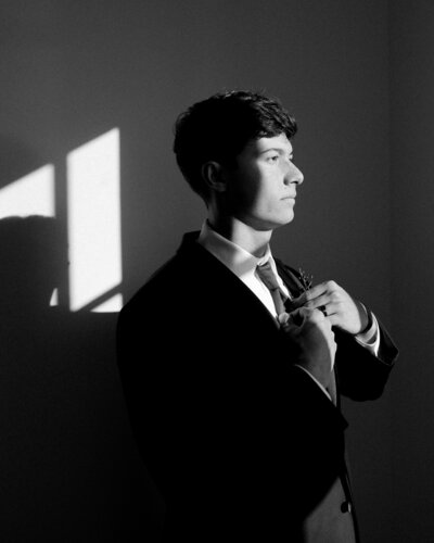 Black and white photo of groom standing against a wall and fixing his tie