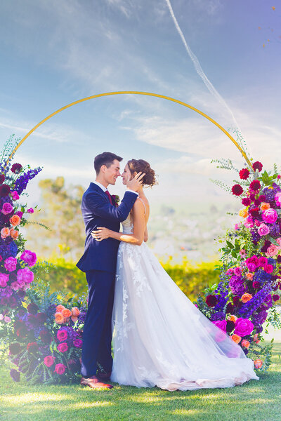 The circle wedding alter is glowing bright with purple, pink and red flowers. The bride and groom say yes in front of the flowered alter by Primrose and Petals.