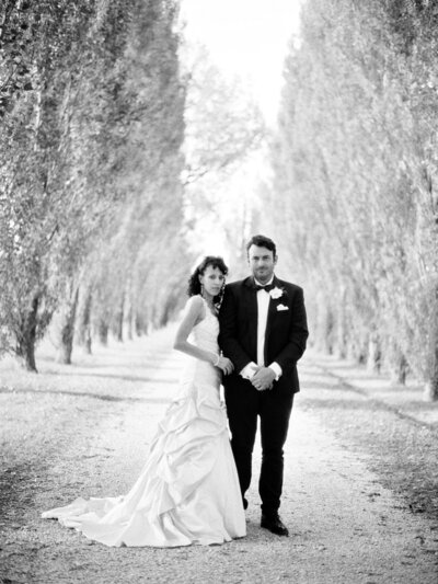 Bride and groom stand together in tree lined path
