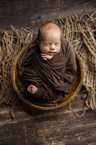 newborn baby wrapped in brown fabric asleep in a wooden bowl