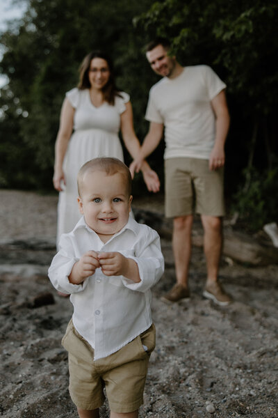 Family session captured by Morgan Ashley Lynn Photography on Lake Michigan in Wisconsin with mom and dad standing in the background looking at toddler son and smiling, toddler is in foreground holding sand and smiling looking directly at the camera