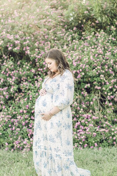 Expecting mom gazing and holding baby bump in front of floral greenery at The Meadow in  Mesa, AZ
