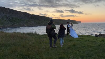 Two photographers capture a bride and groom during a retreat. The sunset elopement by a coastal cliff, evoking the spirit of an adventure elopement with emotional vows.