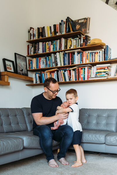 Newborn Photographer, older sibling gives new baby a hug with dad in the living room