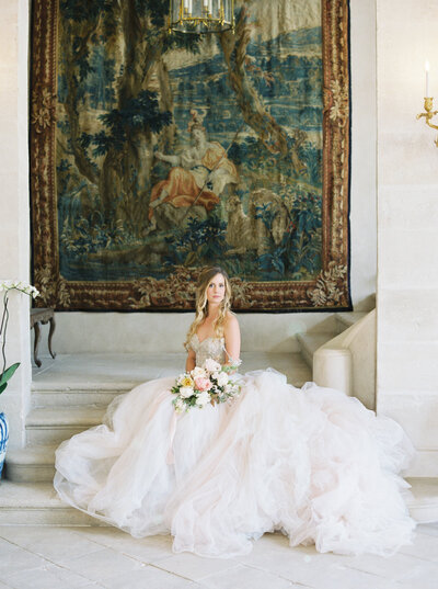 A bride sits under a large tapestry.