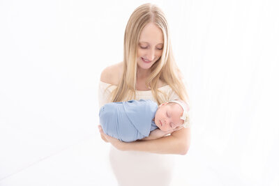 A blonde mother cradling her sleeping newborn baby in a tight blue swaddle while standing in a studio