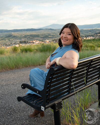 Photo of Ashlyn sitting on a bench in a blue denim dress and looking over her left shoulder at the camera. Mountains and city in the background