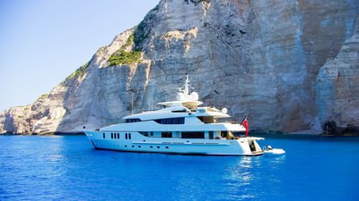 A luxurious white yacht floats on clear blue water against a backdrop of towering rocky cliffs. The vibrant sky reflects on the sea, enhancing the serene and picturesque coastal scene.