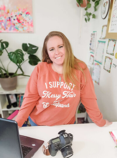 Waman stands with hands on desk wearing sweatshirt that reads "I support messy hair and sweatpants."