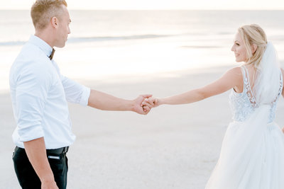 Bride leads her groom by the hand and looks back lovingly on a beach