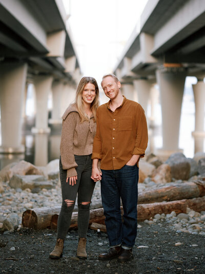 Couple  stands holding hands and smile under a Seattle bridge during an engagement photoshoot