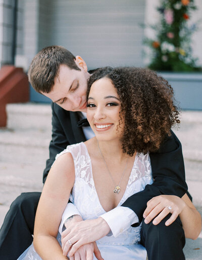 stunning bride in a white lace jumpsuit smiling at the camera as her groom kisses her as they sit on the stairs of the Orlo
