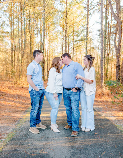 family of four posing standing closely on a road in the woods backlit by golden hour