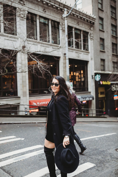 Brunette woman with over the knee boots, dressed in al black with a raybans on and a black pea coat waking across a street in downtown Seattle washinton