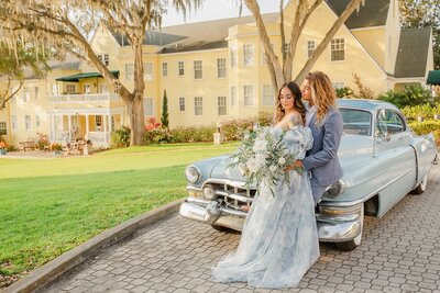 Couple wearing wedding attire posed with a powder blue vintage car in front of a buttercream yellow building