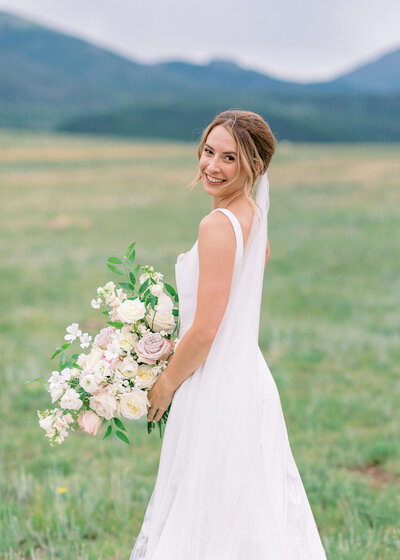 Bride stands in a field smiling at the camera while holding her bouquet of flowers