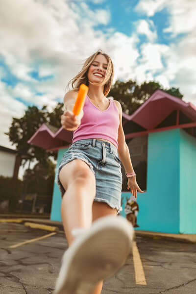 Senior girl wearing a pink tank top and cut off blue jean shorts playfully kicks at camera while eating an orange popsicle