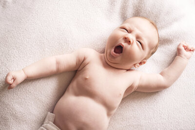 Ein Neugeborenes Baby liegt mit dem Rücken auf einer Decke, gähnt genüsslich und streckt sich.