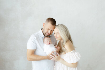 Photo of mom and dad holding a newborn baby girl as they lean in to each other and look at her as they stand in a Dallas photography studio for their baby girls newborn photos.
