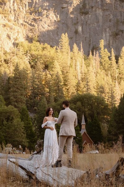 A couple shares a moment during their California elopement, standing on a fallen tree in a golden meadow surrounded by tall pines, with a small rustic chapel and granite cliffs in the background in Yosemite National Park.