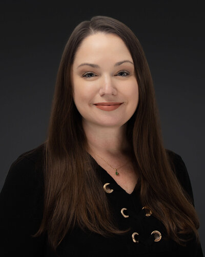 A person stands on stone steps in front of a large stone building, holding a bright red coat. They are wearing a white dress and red shoes. Trees are visible in the background.