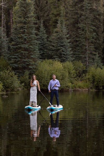 A bride in a white dress and a groom  in a purple shirt stand on paddle boards in the middle of a lake in Idaho.