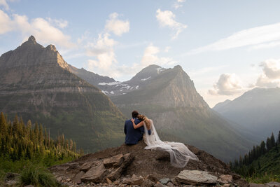 A bride rests her head on her grooms shoulder as they sit on a rock watching the sunset.