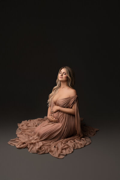 Woman poses for fine art maternity photos in Philadelphia.  She is sitting on her knees angled away from the camera. She is wearing an off-the-shoulder dusty rose gown with her cleavage exposed. The shoulder of the gown drapes along side of her and the gown is puddled around her. The woman is gently holding her bump. The woman's eyes are closed and slightly angled up toward the light. Captured by best Philadelphia maternity photographer Katie Marshall.