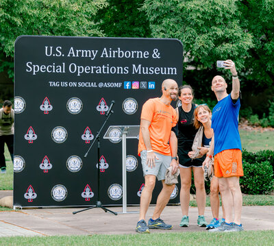 Four people pose for a selfie at the U.S. Army Airborne &amp; Special Operations Museum, with its sign encouraging social media tags. Outside, trees frame the moment beautifully—an ideal shot for any event photographer in Fayetteville, NC.