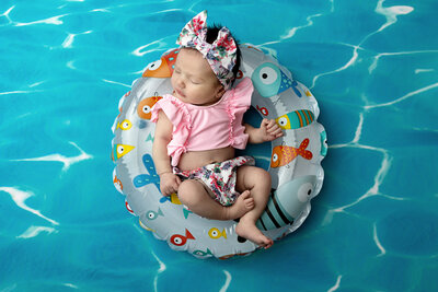 Newborn baby girl wearing a pink & floral bikini asleep in an inner tube on a pool water background
