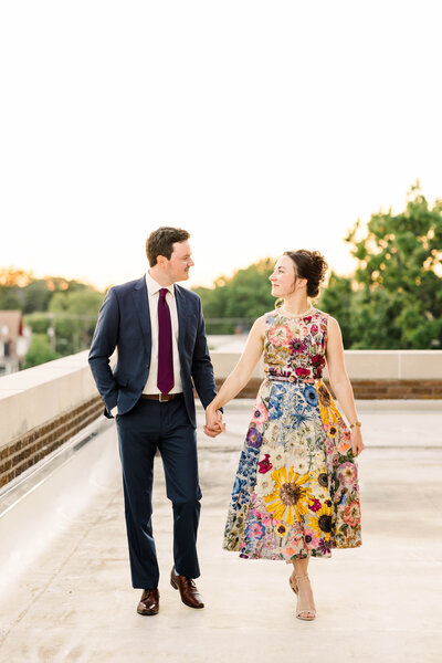 Bride and groom holding hands while walking towards the camera during couple portraits on a balcony overlooking the sunset. The bride is wearing a wedding dress covered in brightly embroidered flowers which highlights Kansas by Kansas City Wedding Photographer Sarah Riner Photography.
