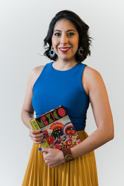 The visionary behind Soleil Vida Studio! The owner, dressed in a vibrant blue top, long yellow skirt, and stylish accessories, stands against a white wall. Short, curled hair frames her face, complemented by long blue earrings and a chic brown watch. In her hand, a book with a colorful cover bearing the word 'Mexico' hints at the cultural richness that inspires her craft.