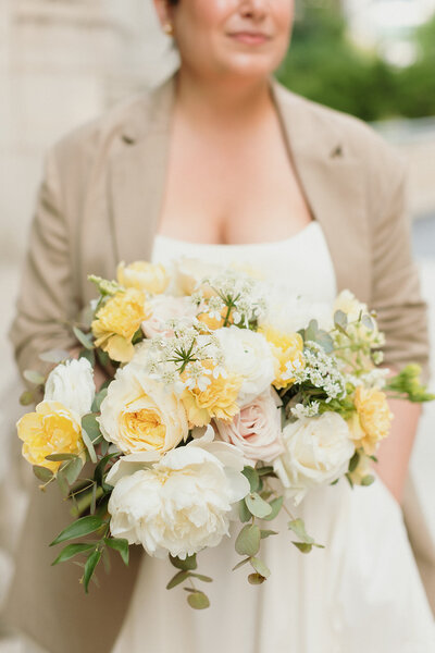 fair skinned bride in white dress and tan jacket hold large bouquet of yellow and white flowers