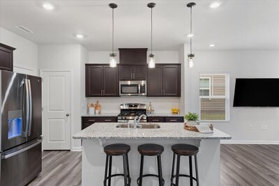  A sleek kitchen with dark wood cabinets, stainless steel appliances, granite countertops, a center island with sink and three barstools, pendant lights overhead, and modern vinyl plank flooring.